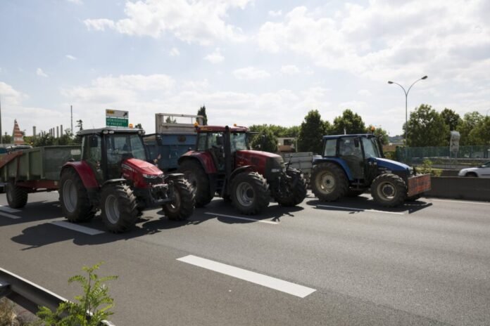 Trois tracteurs agricoles bloquant une autoroute lors d'une manifestation, avec un ciel partiellement nuageux et des éléments urbains en arrière-plan.
