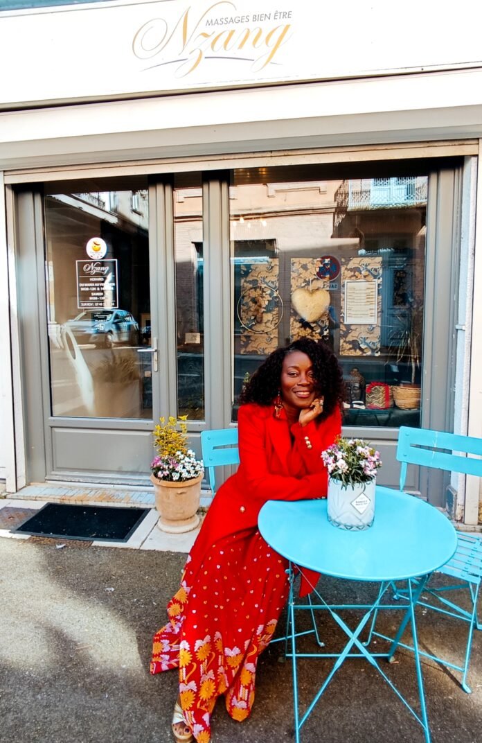 Marlyne, propriétaire de Nzang Massage, assise devant son salon dans une tenue rouge vif